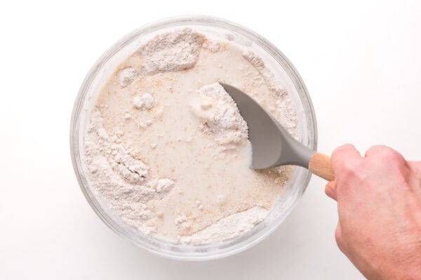A hand holds a spatula, stirring flour with milk ingredients in a glass mixing bowl.