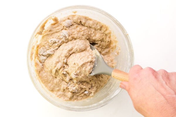 A hand holds a spatula, stirring donut batter in a glass bowl.