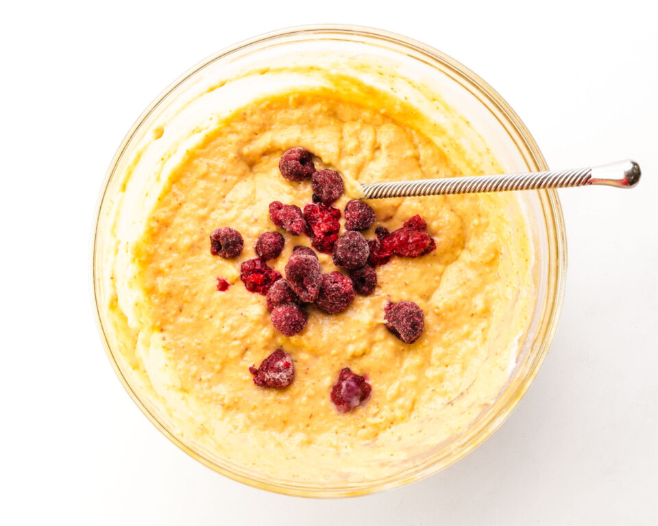 Looking down on a bowl of muffin batter with raspberries in the center. There is a spoon for stirring in the batter too.