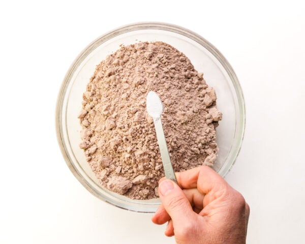 A hand holds a measuring spoon of baking powder over a bowl with boxed brownie mix in it.