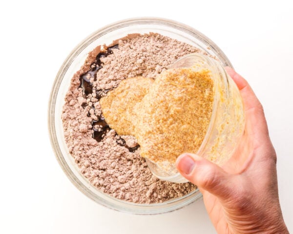 A hand pours a faux egg mixture into a bowl with brownie mix and vegetable oil.