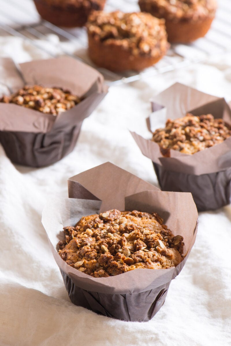 Several coffee cake muffins in papers in the foreground with more muffins on a cooling rack in the background.