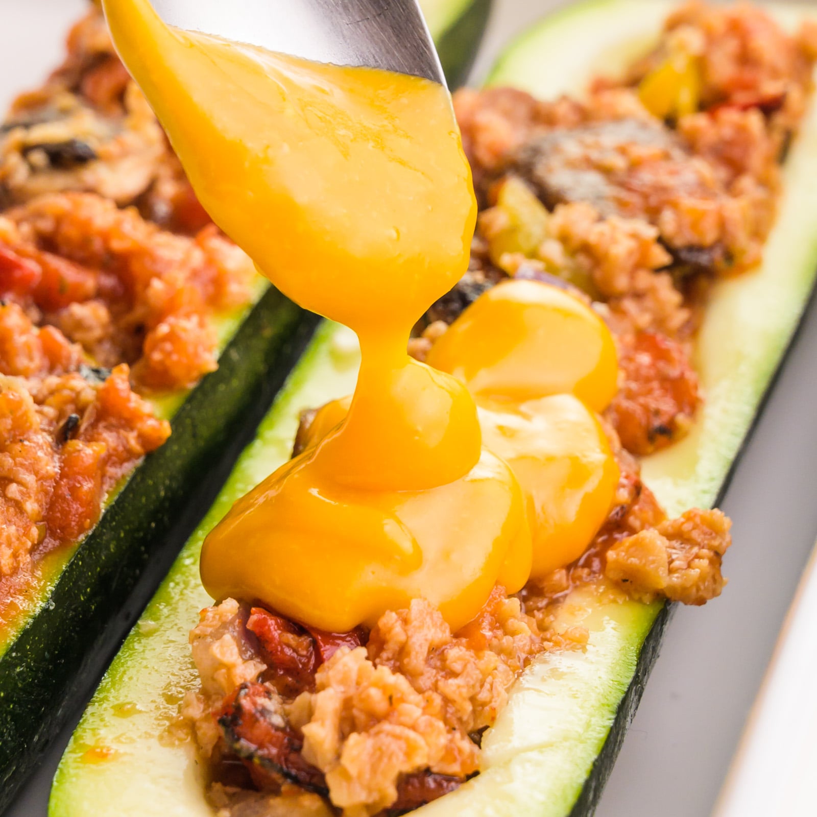 A spoon drizzles melty vegan cheese over zucchini boats.
