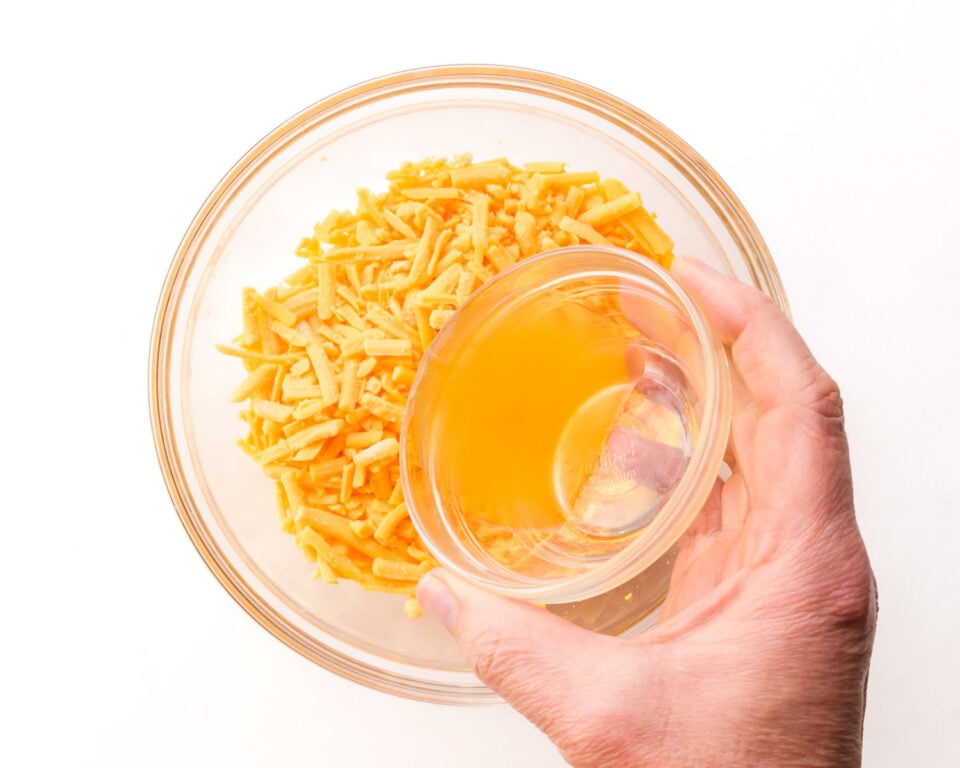 A hand holds a bowl of vegetable broth, pouring it into a bowl with vegan cheddar shreds.