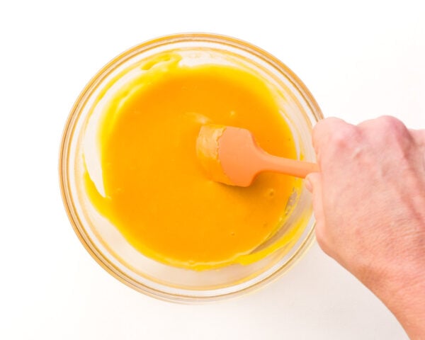 A hand holds a rubber spatula, stirring melted vegan cheese in a bowl.