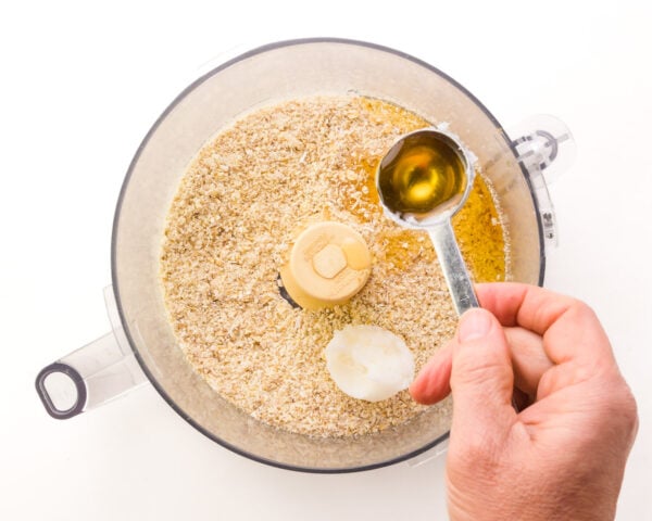 A hand pours agave nectar from a measuring spoon into a food processor bowl with other ingredients, including a scoop of coconut oil.