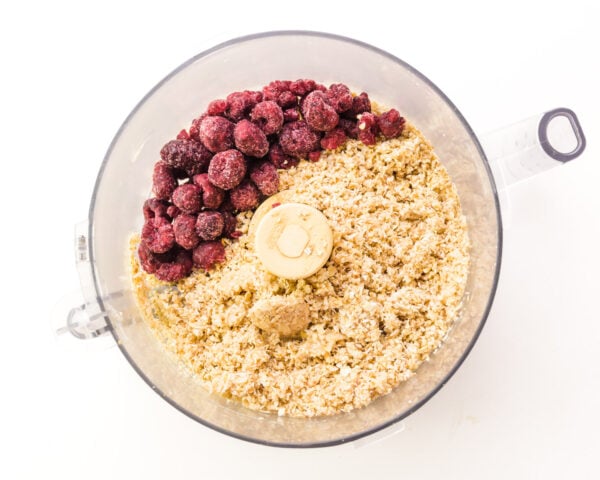 Frozen raspberries are in a food processor along with a mixture of oats and other ingredients that have been ground to a fine texture.