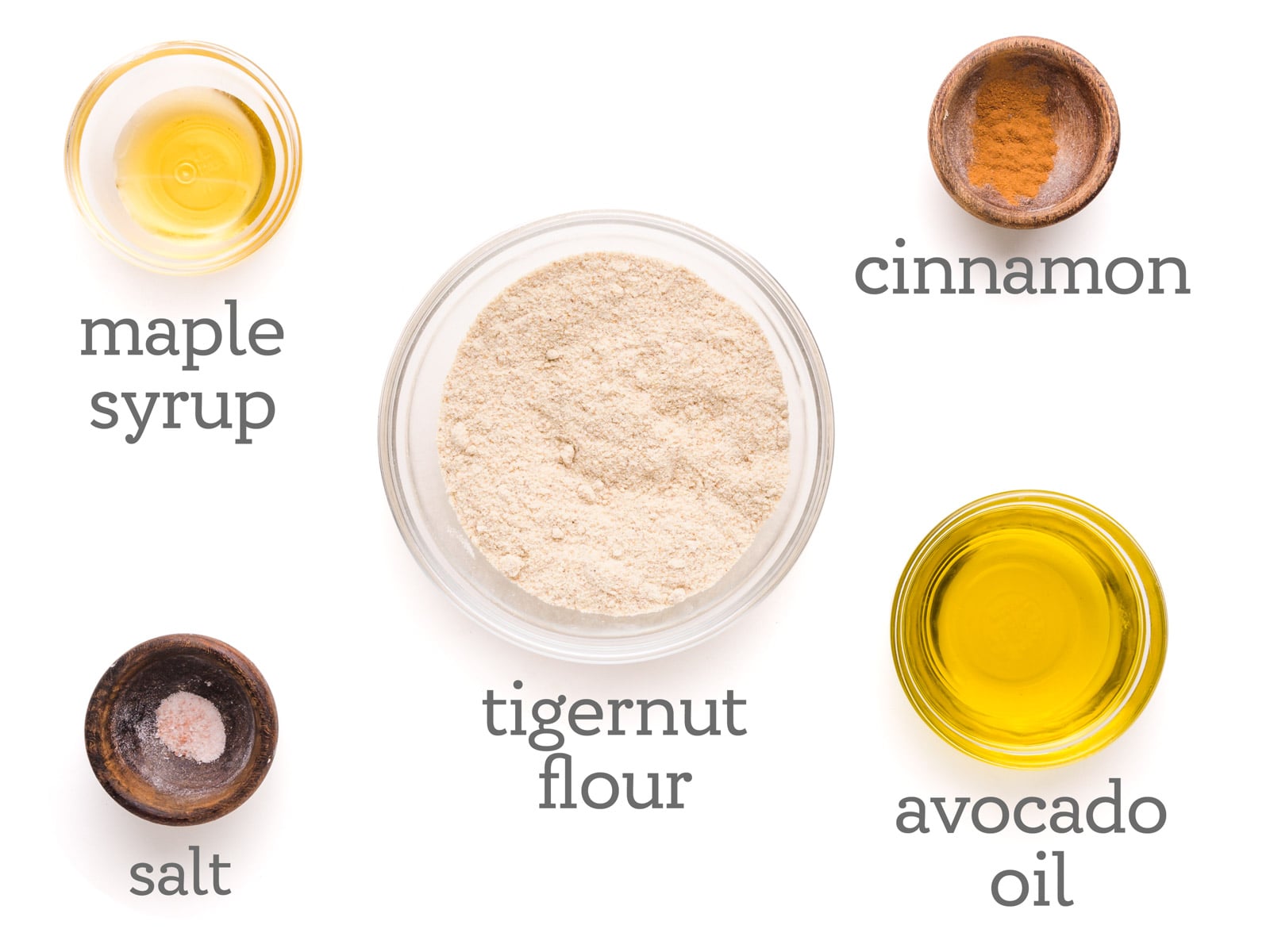 Ingredients in bowls are laid out on a table. The text reads, cinnamon, avocado oil, tigernut flour, salt, and maple syrup.