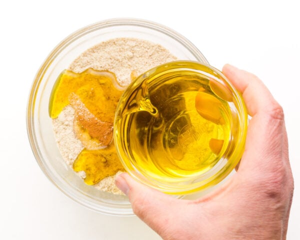 A hand holds a bowl of vegetable oil, pouring it into a bowl with flour.