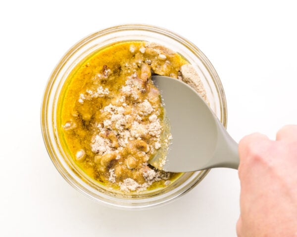 A hand holds a spatula stirring together a mixture of flour and oil in a bowl.