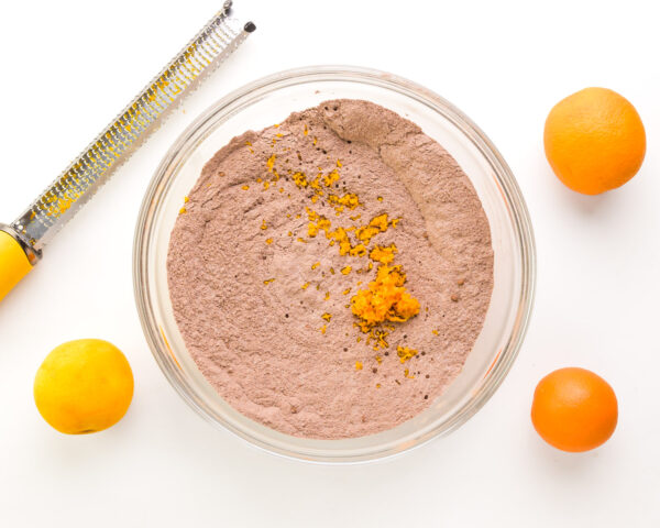 Orange zest is sitting on top of a cocoa flour mixture in a mixing bowl. There are oranges around the bowl and a microplane zester.