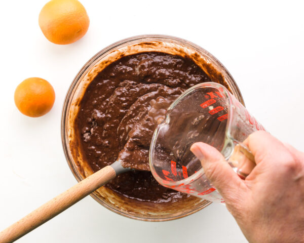 A hand holds a pyrex measuring cup, pouring hot water into chocolate batter in a mixing bowl. There is a spoon in the mixing bowl and two oranges sit next to the bowl.
