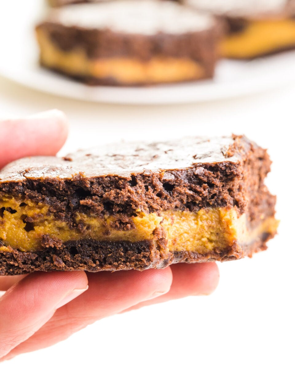 A hand holds a vegan pumpkin brownie with a bite taken out. There are more brownies on a plate in the background.