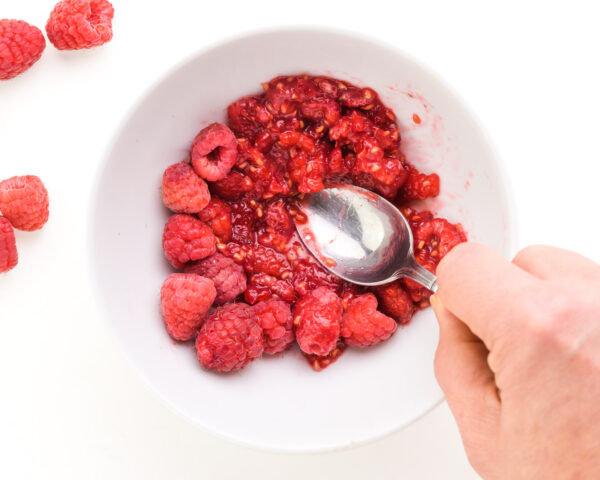 A hand holds a spoon, mashing raspberries in a bowl. There are fresh raspberries beside the bowl.
