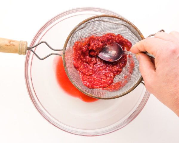A hand pushes mashed raspberries through a fine mesh strainer into a bowl.