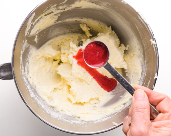 A hand holds a spoonful of mashed raspberries, pouring it over whipped butter in a mixing bowl.