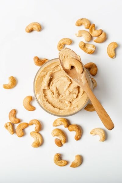 Looking down on a jar of cashew butter. There is a wooden spoon with cashew butter and a cashew. There are several cashews around the jar.