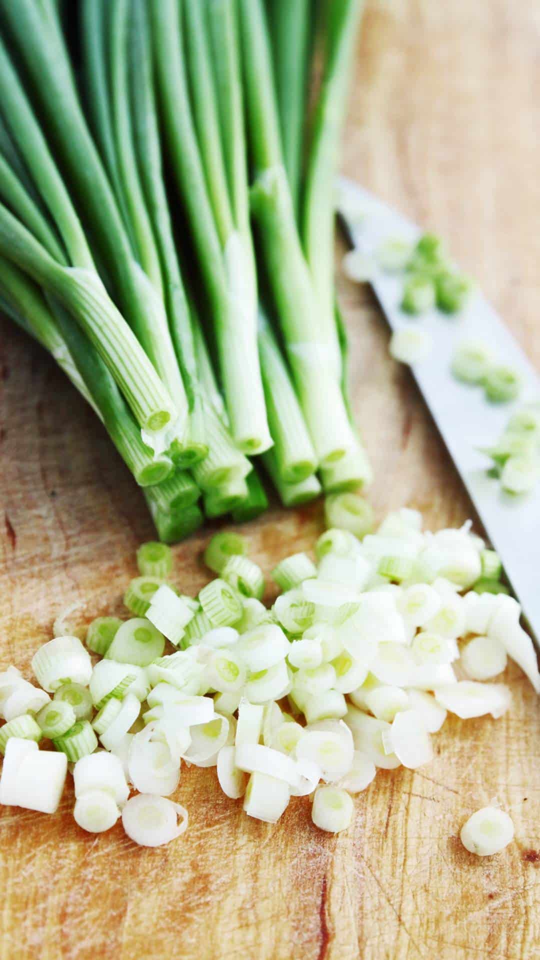 A bunch of spring onions are on a cutting board with some of the white parts of the onion finely chopped. There is a knife beside the onions.