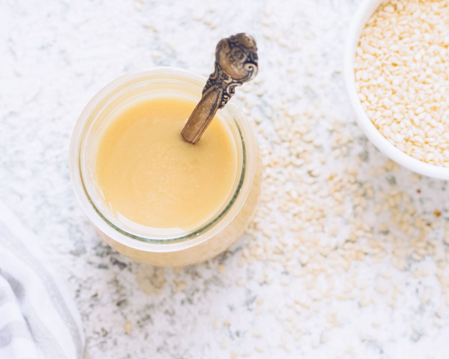 Looking down on a jar of sesame seed butter with a spoon in it. There is a bowl of sesame seeds and more seeds scattered on the table.