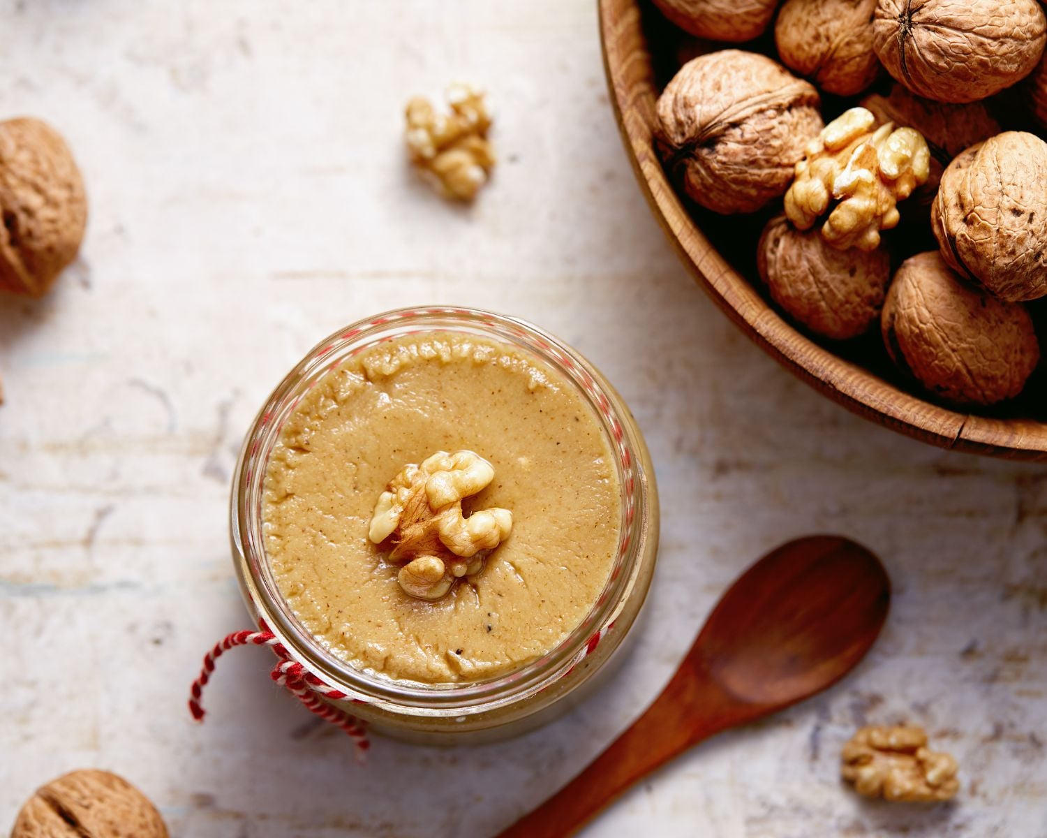 Looking down on nut butter in a jar with a walnut on top. There is a spoon and a bowl of walnuts beside it and more walnuts are scattered around the jar.