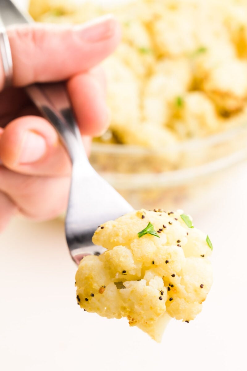 A hand holds a fork with cauliflower on it. The fork hovers in front of a bowl with more cooked cauliflower.