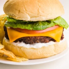 A close-up of an air fryer Impossible Burger on a bun with all the toppings, including cheese and mayo. It sits on a plate next to potato chips.