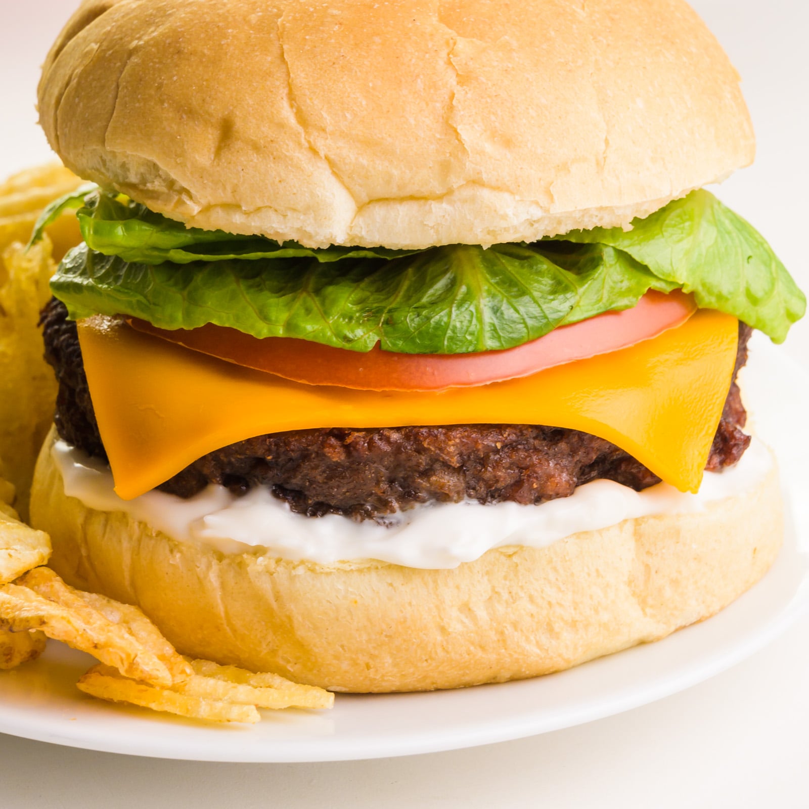 A close-up of an air fryer Impossible Burger on a bun with all the toppings, including cheese and mayo. It sits on a plate next to potato chips.
