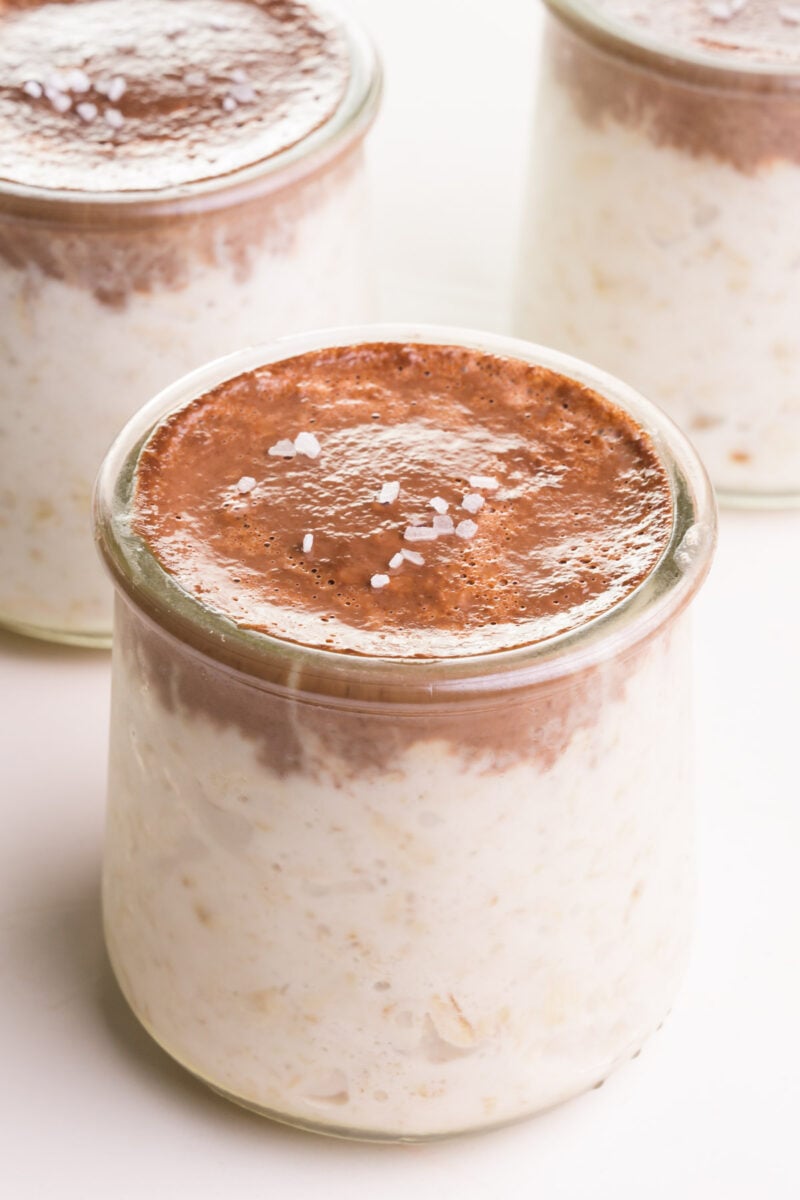 A bowl of bounty overnight oats has a chocolate topping and flaky sea salt on top. There are two jars of the same oats in the background.