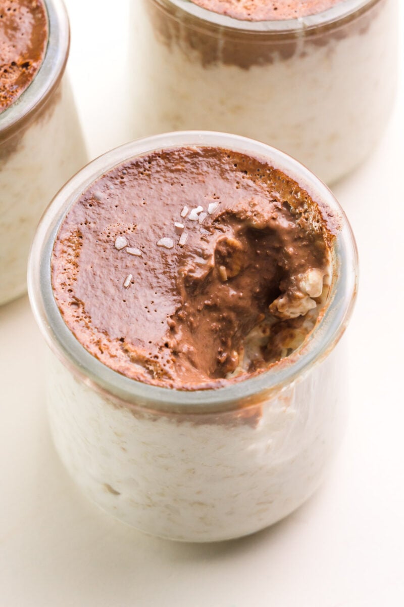 Looking down on a bowl of coconut chocolate overnight oats with a bite taken out. There are two jars with the same oats partially visible in the background.