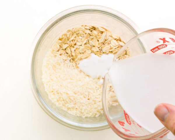A hand holds a bowl of coconut milk, pouring it into another bowl with oats and coconut flakes.