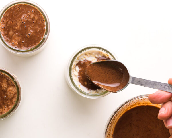 Chocolate sauce is being poured with a spoon over overnight oats.