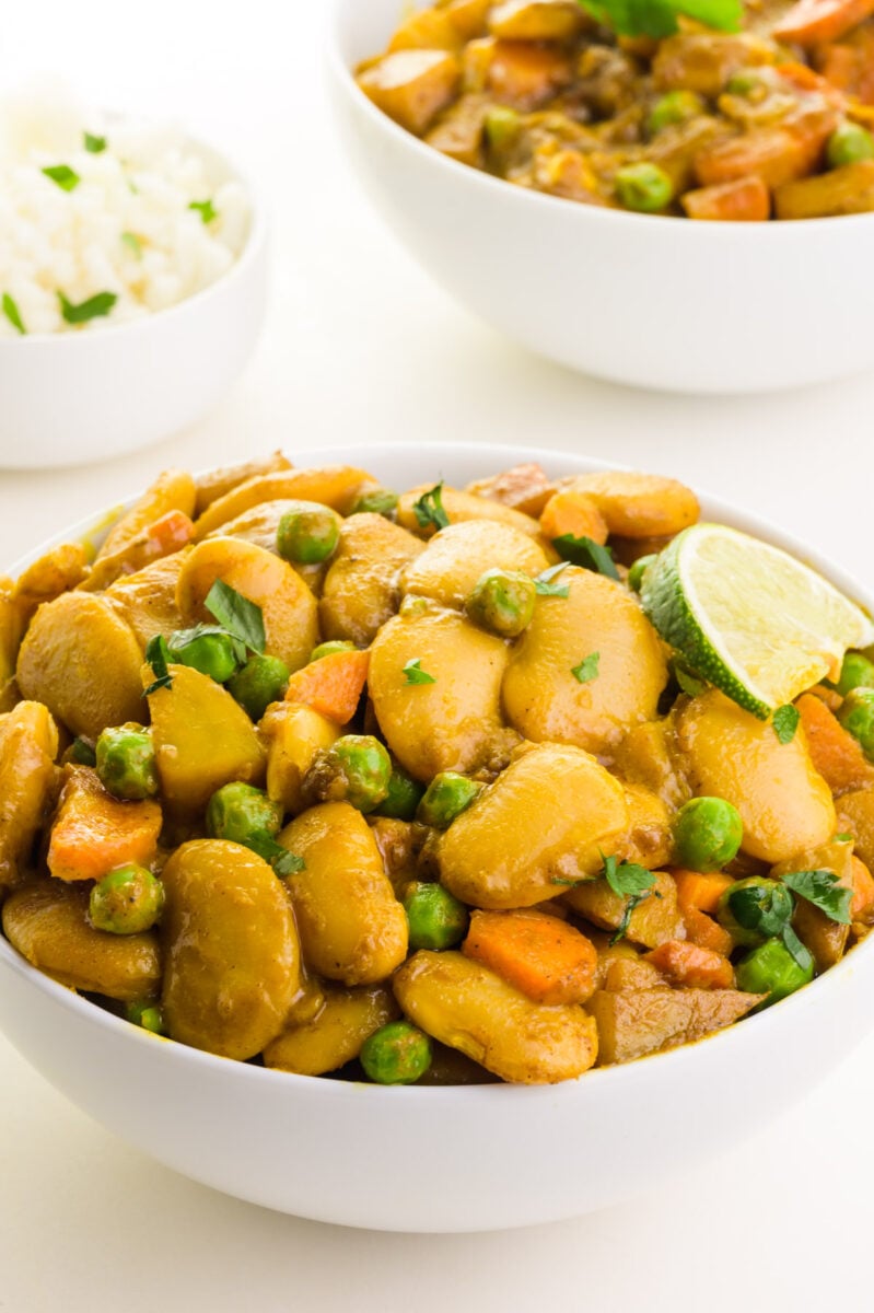 A bowl of curry with butter beans has a slice of lime on the side. There is a bowl of rice and more curry in the background.