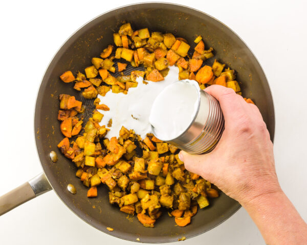 Coconut milk is being poured into a skillet with other ingredients like chopped onions and potatoes.
