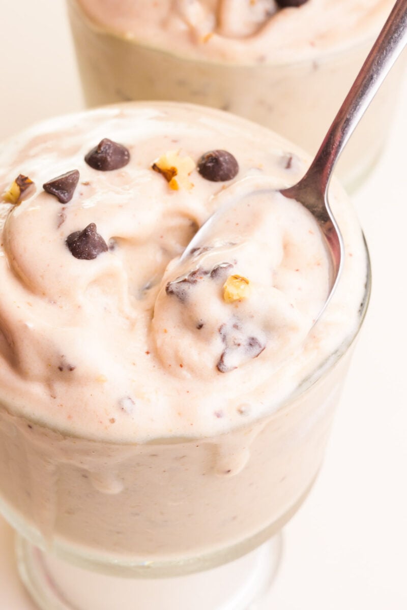 A spon rests in a bowl of vegan chunky monkey ice cream. There are chocolate chips and chopped walnuts on top.