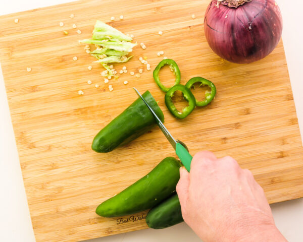 Looking down on a hand holding a paring knife, cutting a jalapeño. There are more jalapeños, the pith and seeds, and a red onion on the cutting board.