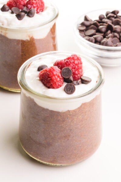 A small glass jar holds chocolate flax pudding topped with creamy white yogurt, chocolate chips an raspberries. There is another jar and a bowl of chocolate chips in the background.
