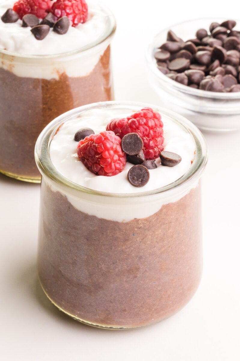 A small glass jar holds chocolate flax pudding topped with creamy white yogurt, chocolate chips an raspberries. There is another jar and a bowl of chocolate chips in the background.
