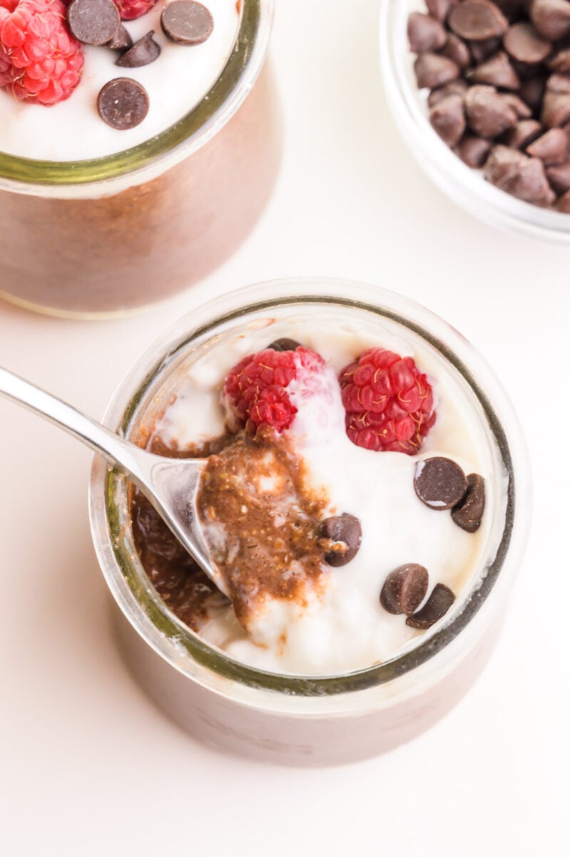Looking down on a jar full of chocolate pudding with yogurt on top and fresh raspberries. There is a spoon, showing some of the chocolate pudding. There is another jar and  bowl of chocolate chips in the background.