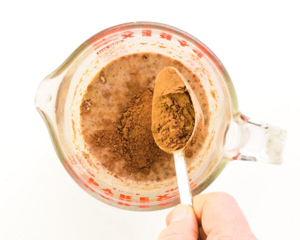 A measuring spoon of cocoa powder is being poured into a pyrex bowl with plant-based milk.
