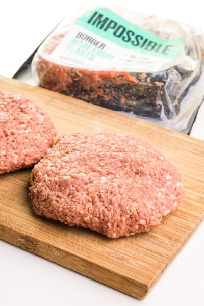 Hand-pressed burger patties are on a wooden cutting board, sitting in front of an opened package of Impossible Burger grounds.