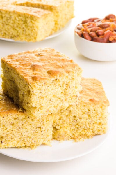 A stack of vegan jiffy cornbread slices sits on a white plate. There is a small bow of chili and a plate with more cornbread in the background.