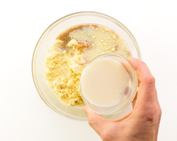 A hand holds a bowl of soy milk, pouring it into a bowl with cornbread ingredients.