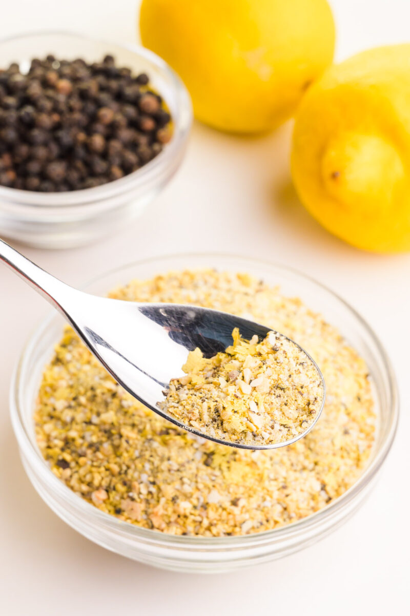 A spoon holds lemon garlic seasoning hovering over a bowl with more of the mixture. There is a bowl of peppercorns in the background along with a couple of fresh lemons.