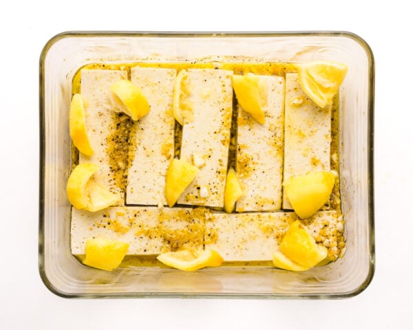 Pieces of tofu are marinading in a shallow glass dish. There are pieces of lemon scattered on top of the tofu.