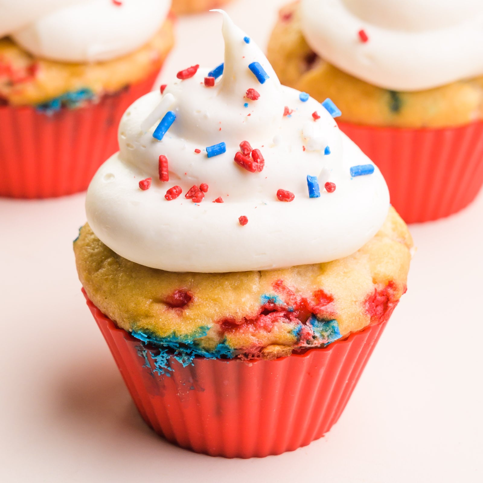 A cupcake in a red silicone cupcake liner has vanilla frosting on top. Both the cupcakes and the frosting are full of red, white, and blue sprinkles. There are more cupcakes in the background.