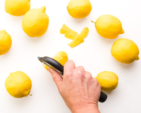 A hand holds a zester, removing the skin from a fresh lemon. There are several fresh lemons around it.