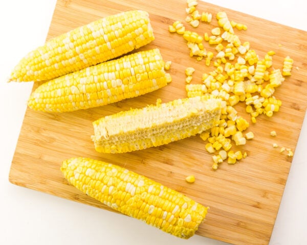 Looking down on ears of corn on a cutting board, one showing some of the husk. There are kernels of corn around them.