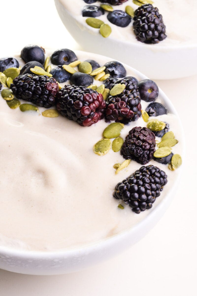 A closeup of a vanilla smoothie bowl with fresh fruit and pumpkin seeds on top. There's another bowl in the background.