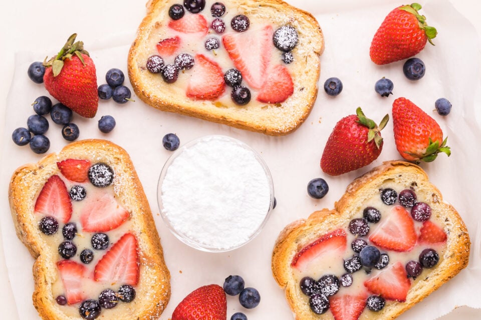 Looking down on several slices of custard toast, sitting next to a bowl of powdered sugar and fresh berries.