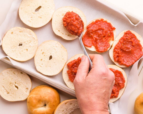 A hand holds a spoon, spreading pizza sauce over mini bagels laid out on a baking pan.